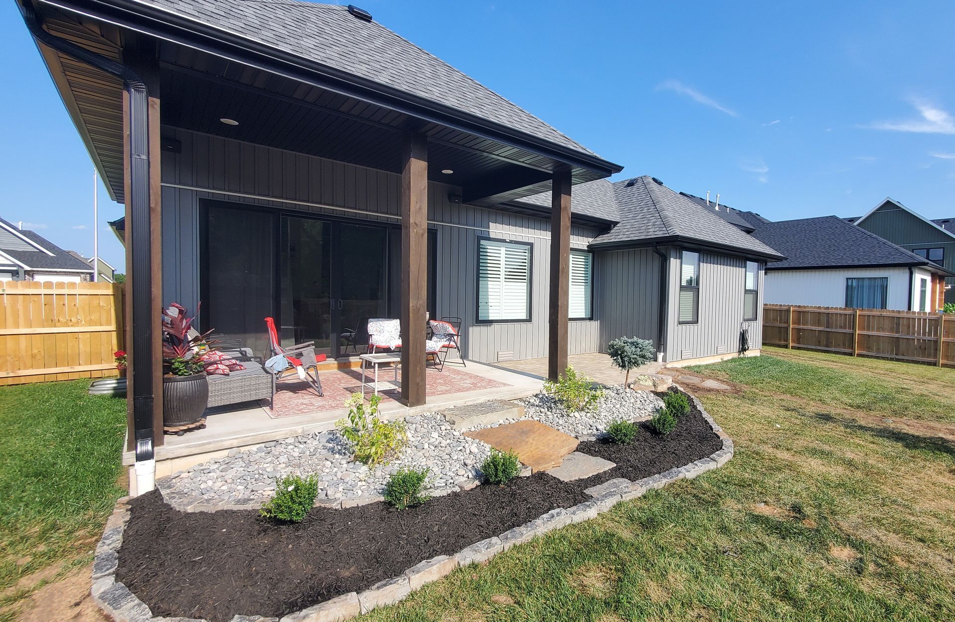 Backyard patio with a gray house, wooden beams, landscaping, and a blue sky.