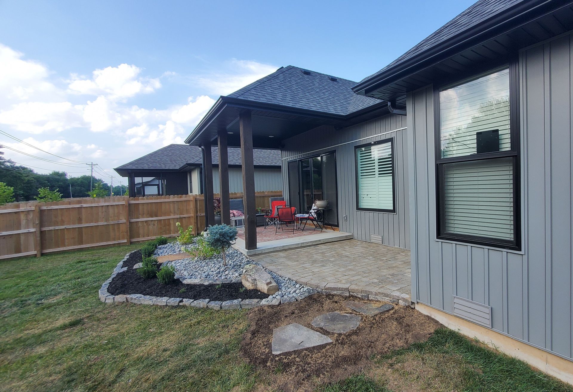 Exterior of a modern house with a patio and landscaping; gray siding, wooden supports, and gravel patio.