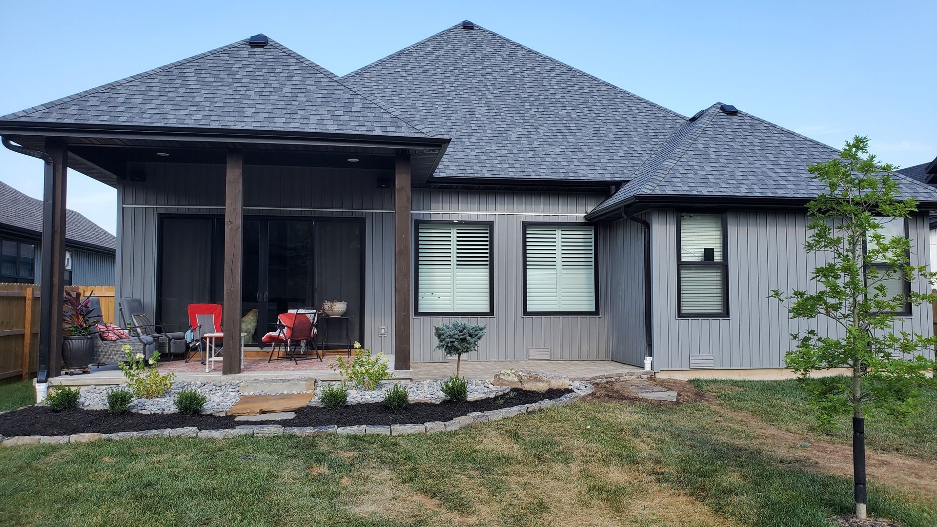 Grey house with dark roof and trim, covered patio, windows, and small yard with grass and landscaping.