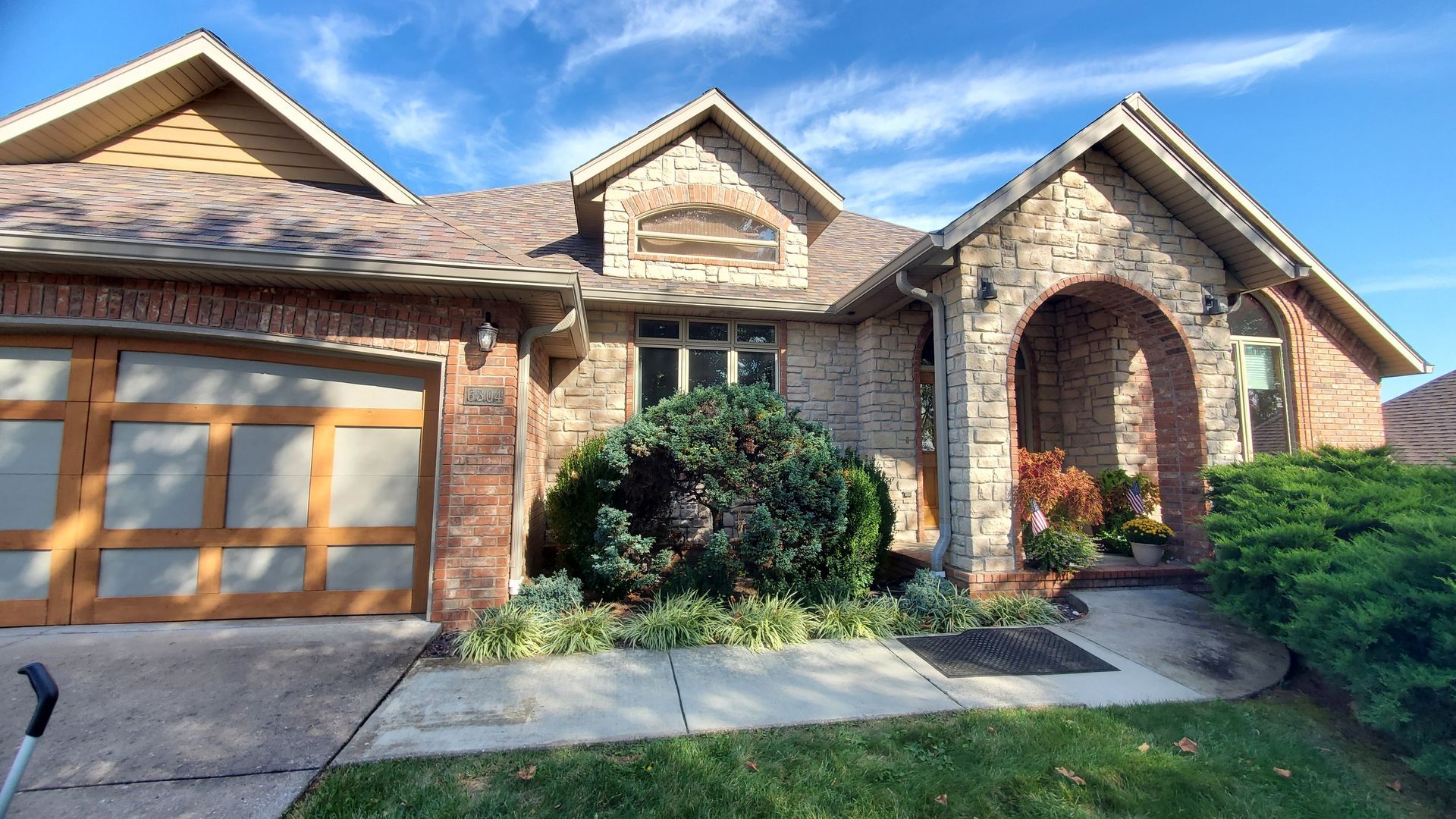 Brick house with arched entry and two-car garage under a blue sky, surrounded by green landscaping.