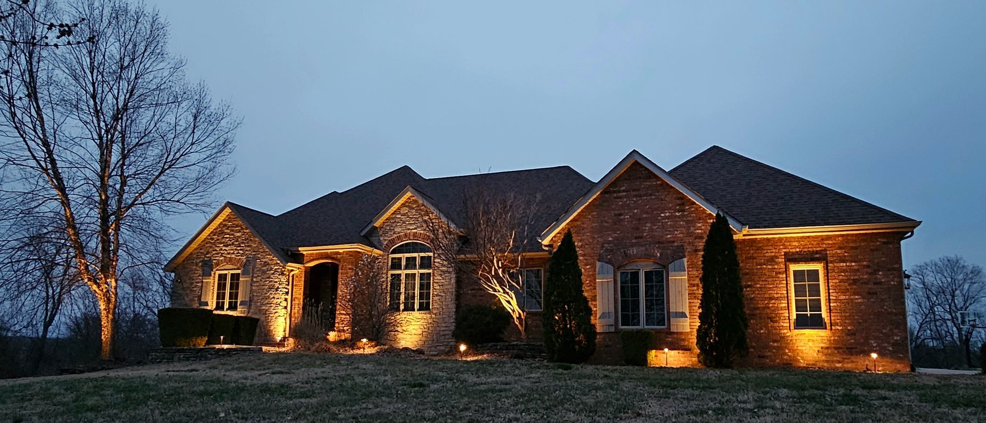 House at dusk, illuminated by outdoor lights, set against a blue sky, and a bare tree.