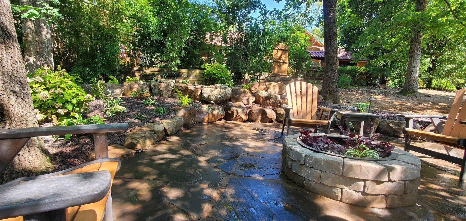 Outdoor patio with fire pit, wooden chairs, and lush greenery. Sunny day.