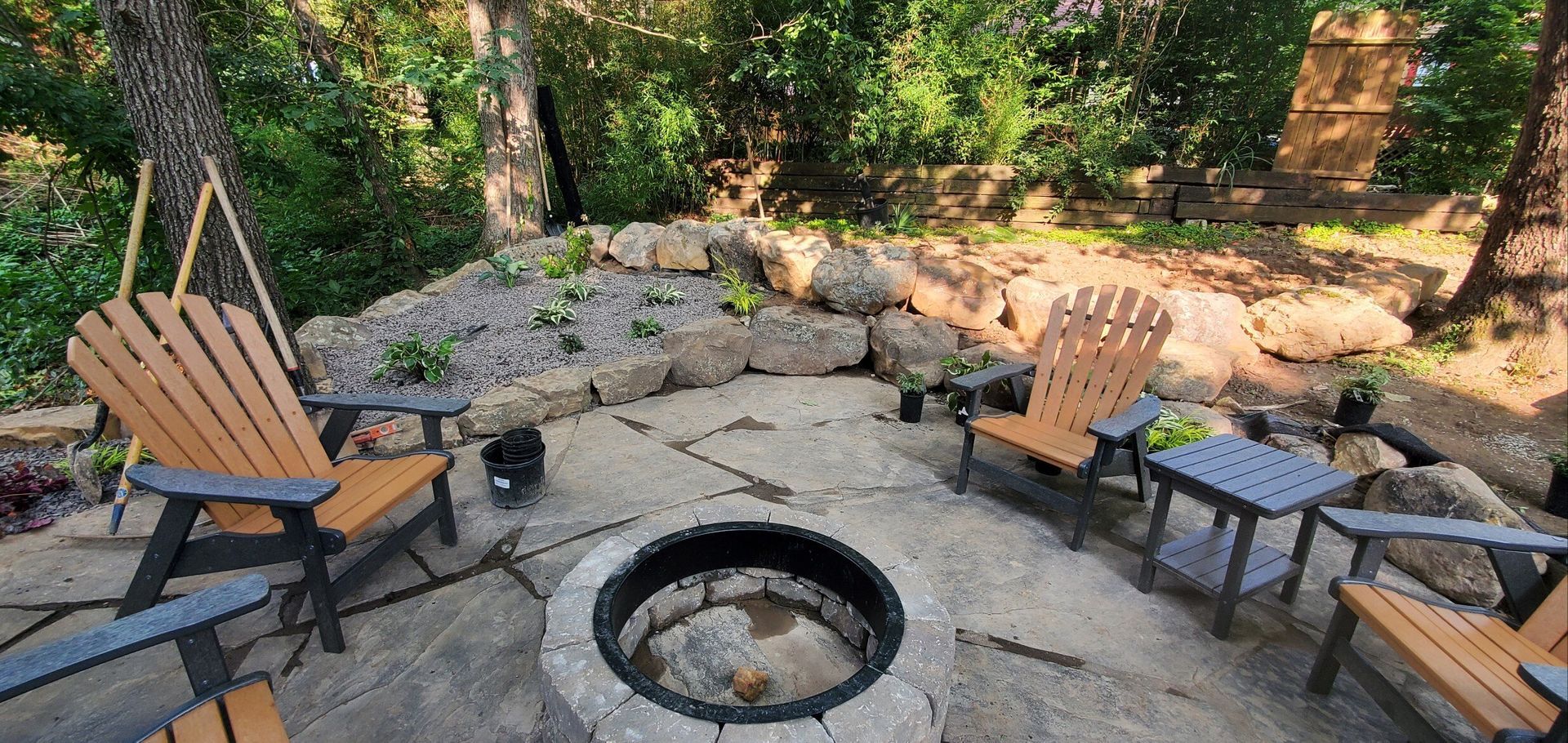 A fire pit surrounded by stone pavers, Adirondack chairs and a stone retaining wall in a backyard setting.