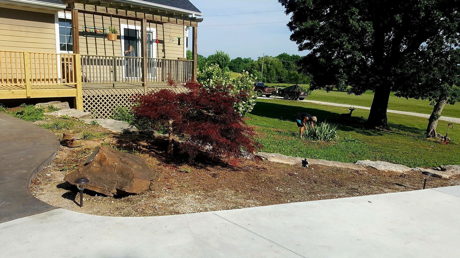 A house with a porch and landscaping, including a red-leafed bush and a large rock, with a green field in the background.