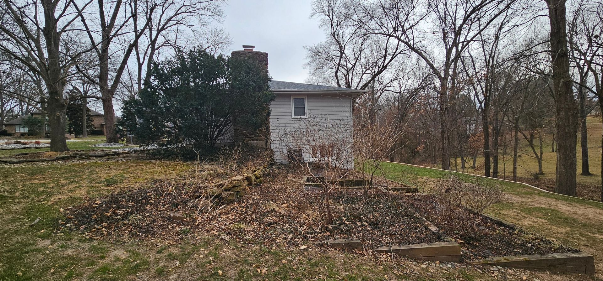 A house with a porch and landscaping, including a red-leafed bush and a large rock, with a green field in the background.