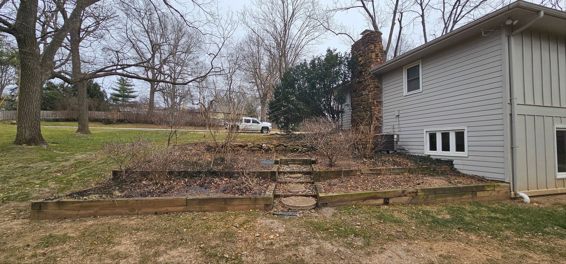 A house with a porch and landscaping, including a red-leafed bush and a large rock, with a green field in the background.