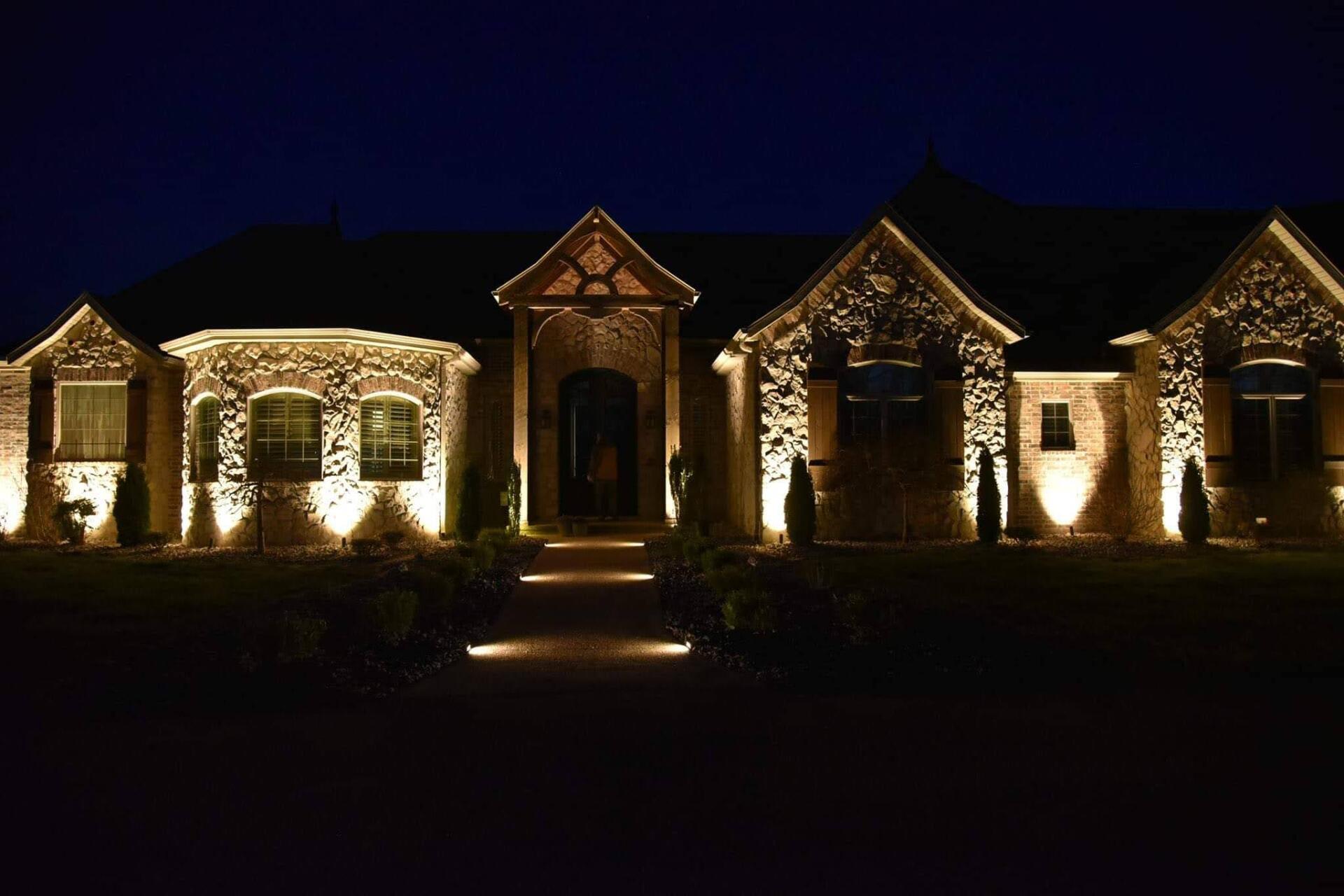 A house illuminated at night with outdoor lighting showcasing stonework and front walkway.