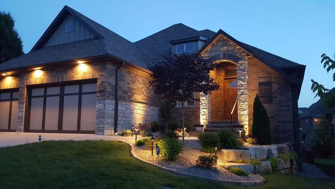 Stone house illuminated at dusk, with a landscaped front yard and dark garage doors.