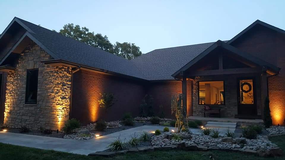 House exterior at dusk with stone facade, warm spotlights, and a pathway leading to the front door.