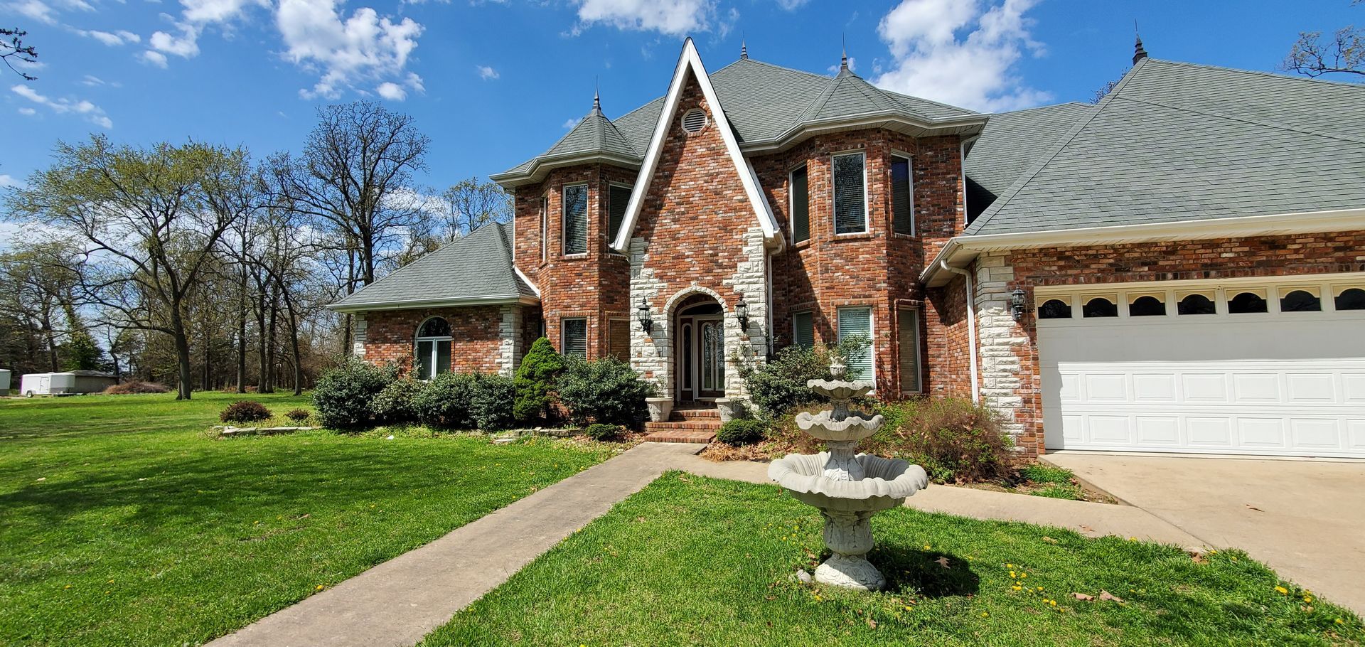 A brick home with a stone roof and a wide driveway. The house is on a large green lawn with trees.