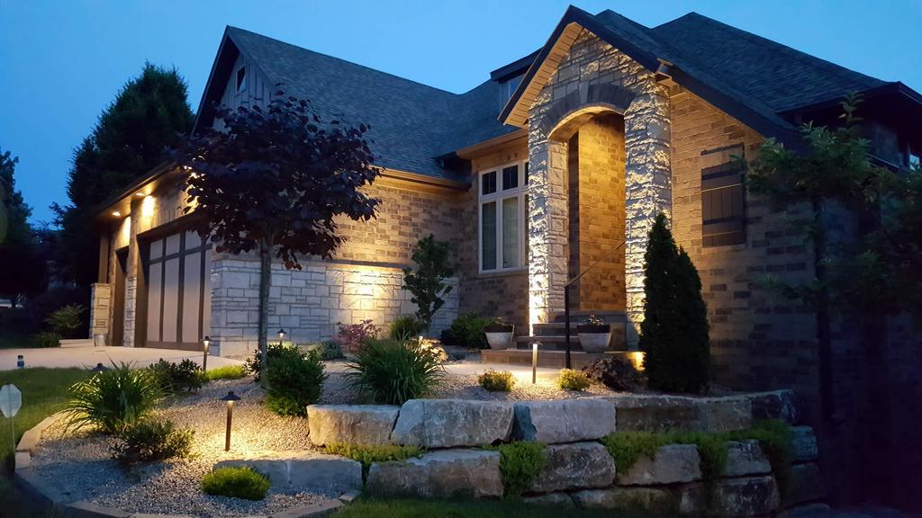 House exterior at dusk, illuminated with landscape lighting highlighting stone facade and landscaping.