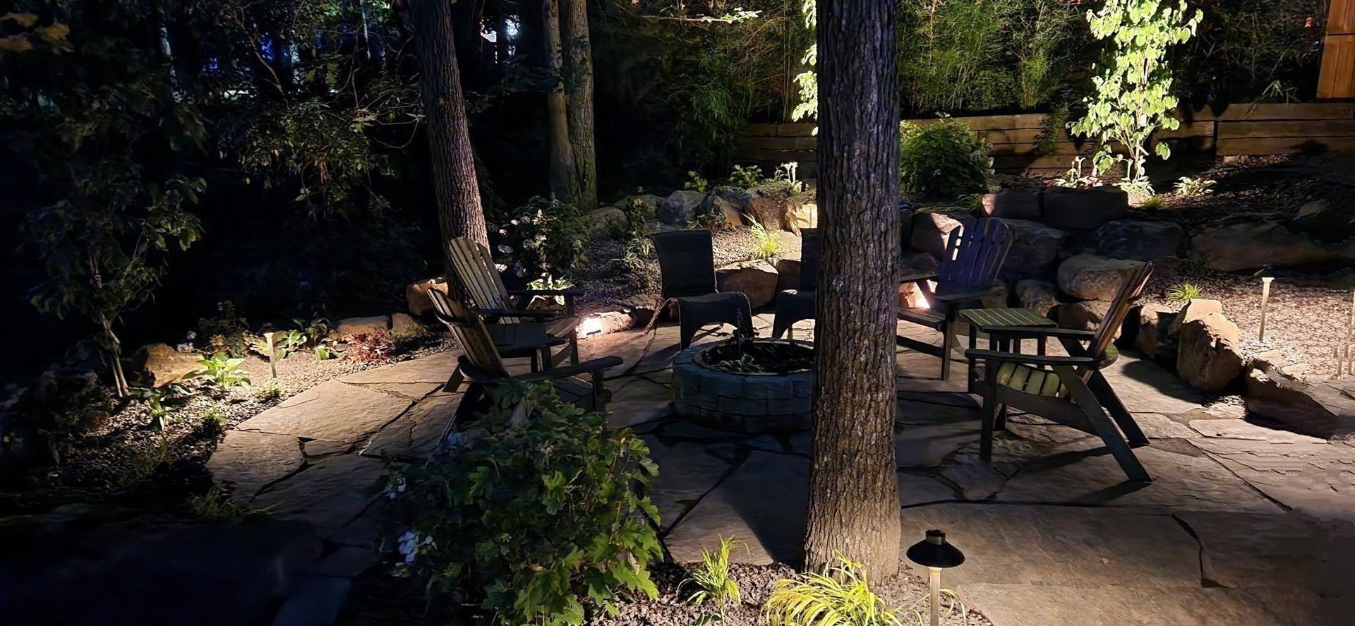 Night view of an outdoor patio with trees and chairs around a fire pit illuminated by spotlights.