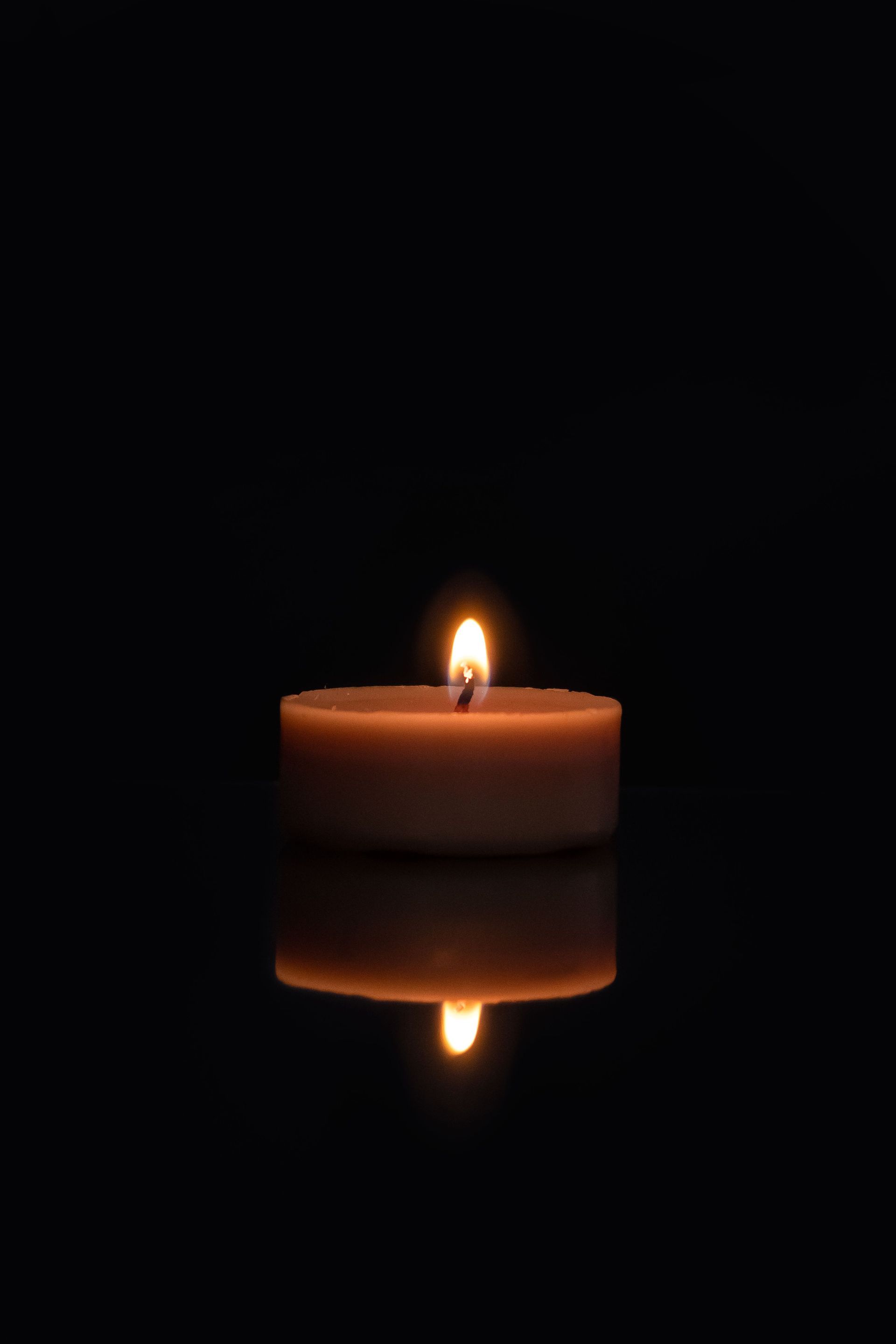 Lit candle reflected on a dark, glossy surface in a black background