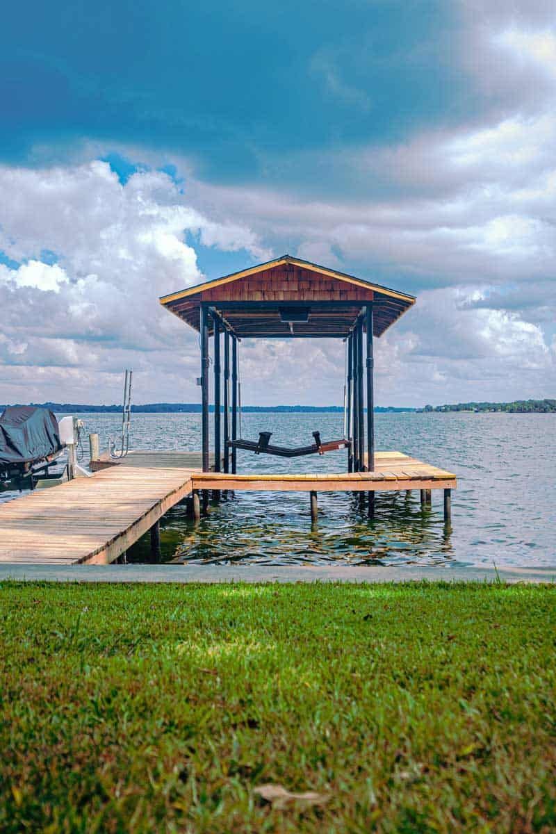 Small lakeside dock with a covered seating area under a cloudy sky, surrounded by water and grass.