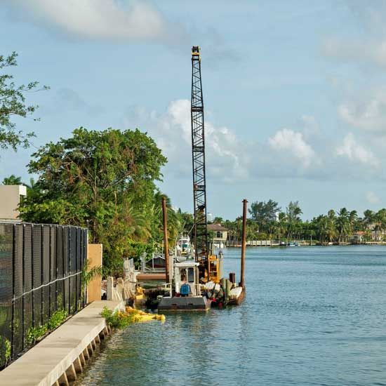 Riverfront construction site with a tall drilling rig beside calm water and palm trees under a blue sky