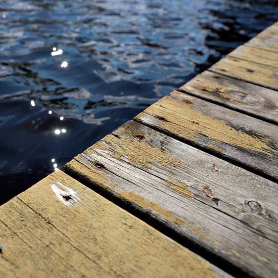 Wooden dock corner next to rippling blue water. Sunlight reflects on the water's surface.