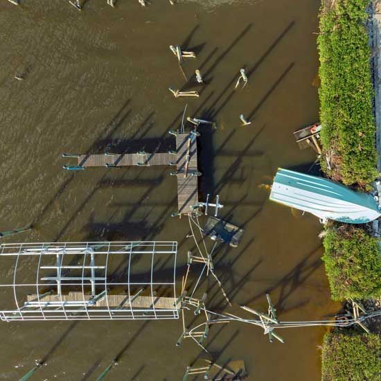 Aerial view of a muddy canal with docks, small boats, and long shadows from mooring posts.