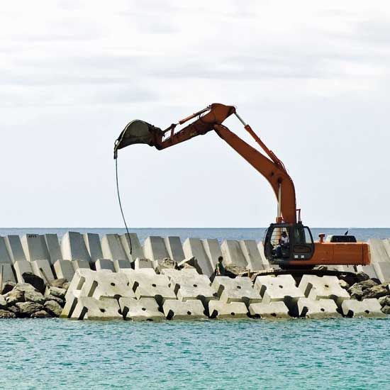 Orange excavator on a rocky seawall lifting debris beside blue water under a cloudy sky