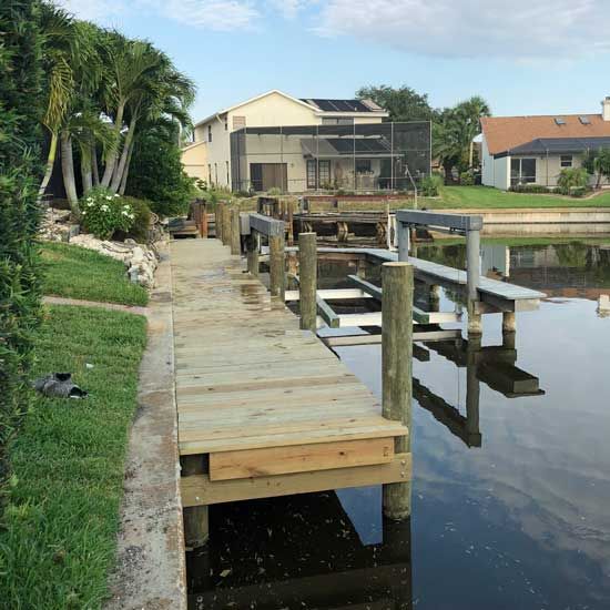Wooden dock beside a calm canal, with houses and palm trees along the shore.