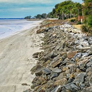 Beach with sand, rocks, and a wooden walkway along the shore under a blue sky.