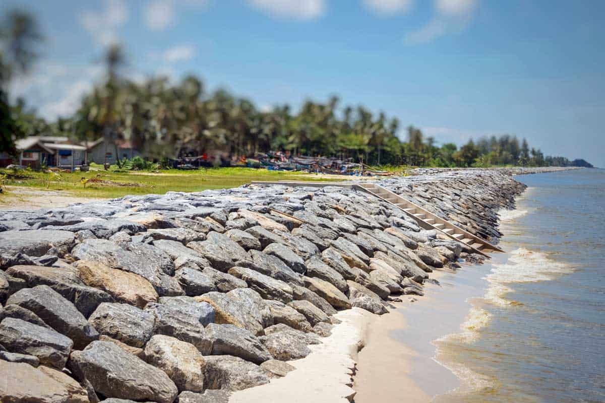 Rocky shoreline with a seawall beside calm water and palm trees under a blue sky
