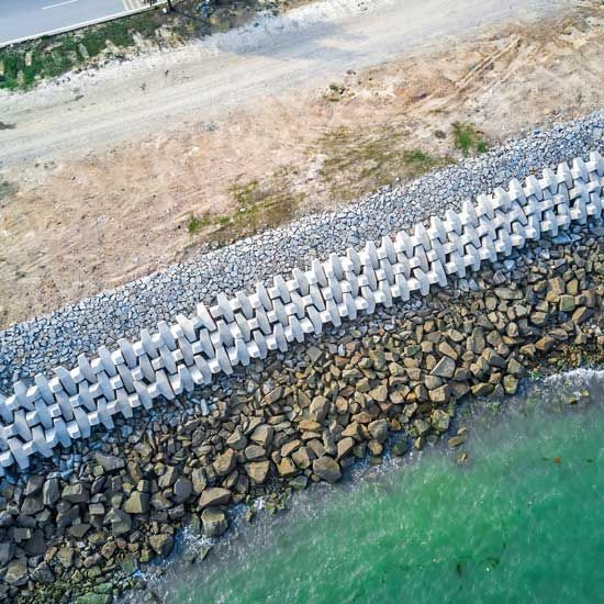 Aerial view of a rocky shoreline with concrete wave breakers along clear turquoise water and a dirt road above.