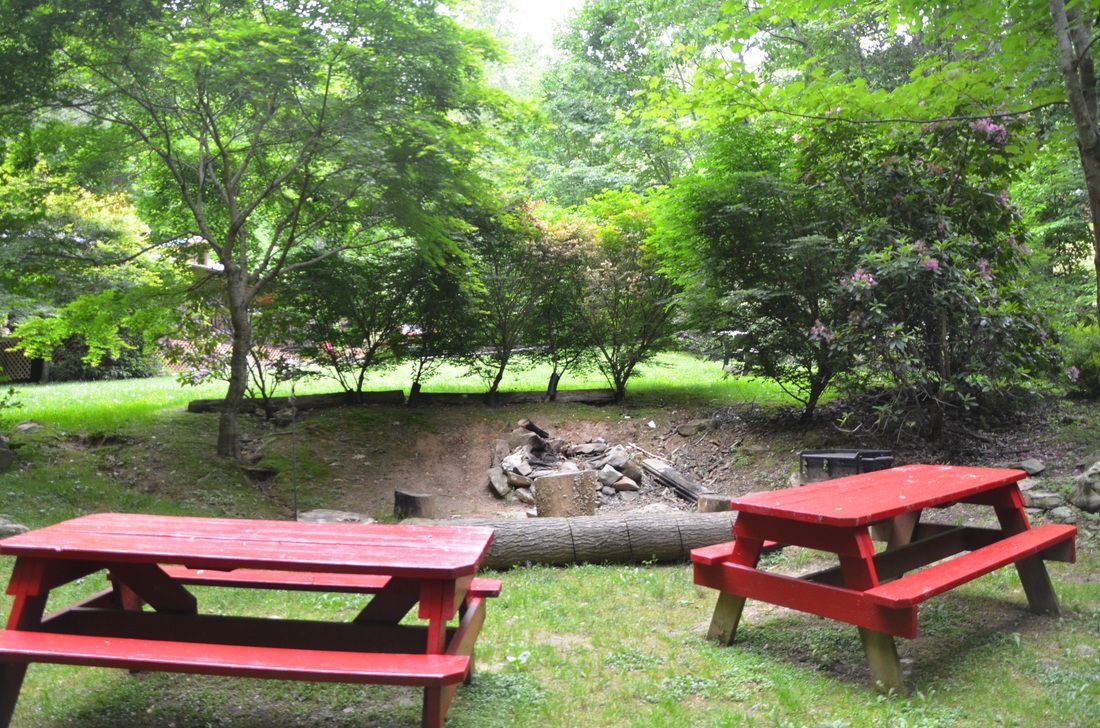Red picnic tables on grass beside a small rock-lined fire pit in a leafy park