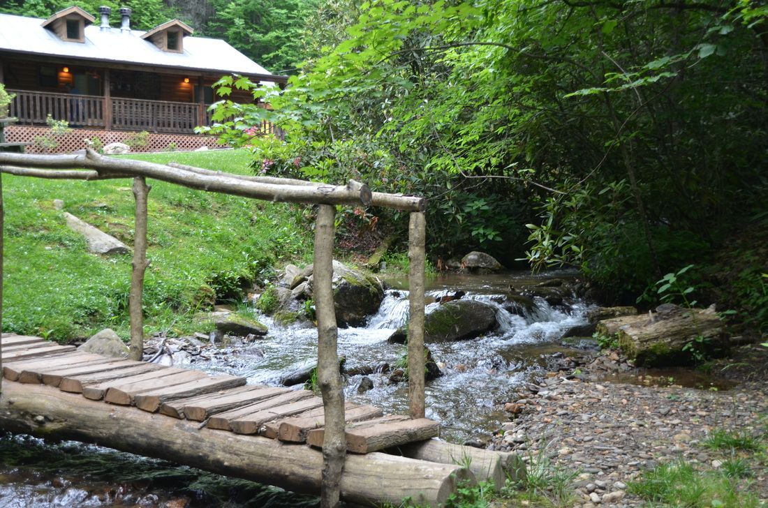Wooden footbridge over a rocky stream in a lush forest near a rustic cabin