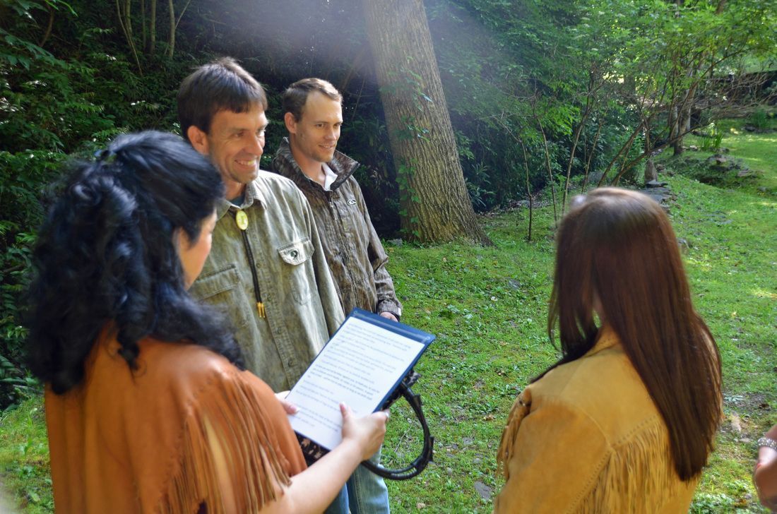 Four people stand outdoors in a garden, one holding a paper while facing two others.