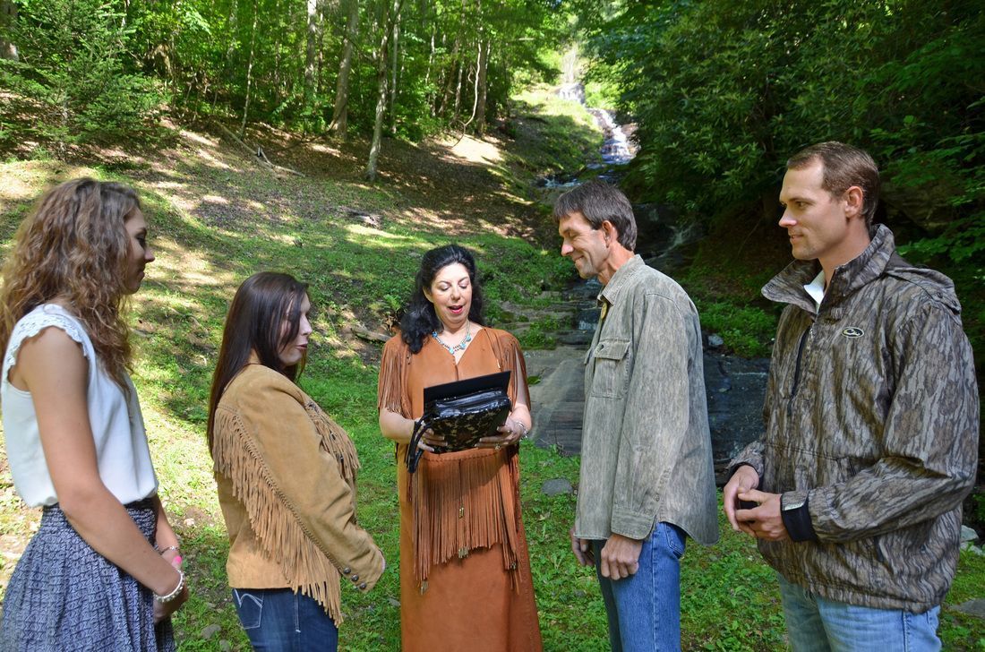 Five people stand outdoors in a wooded area, one holding a laptop while others listen.
