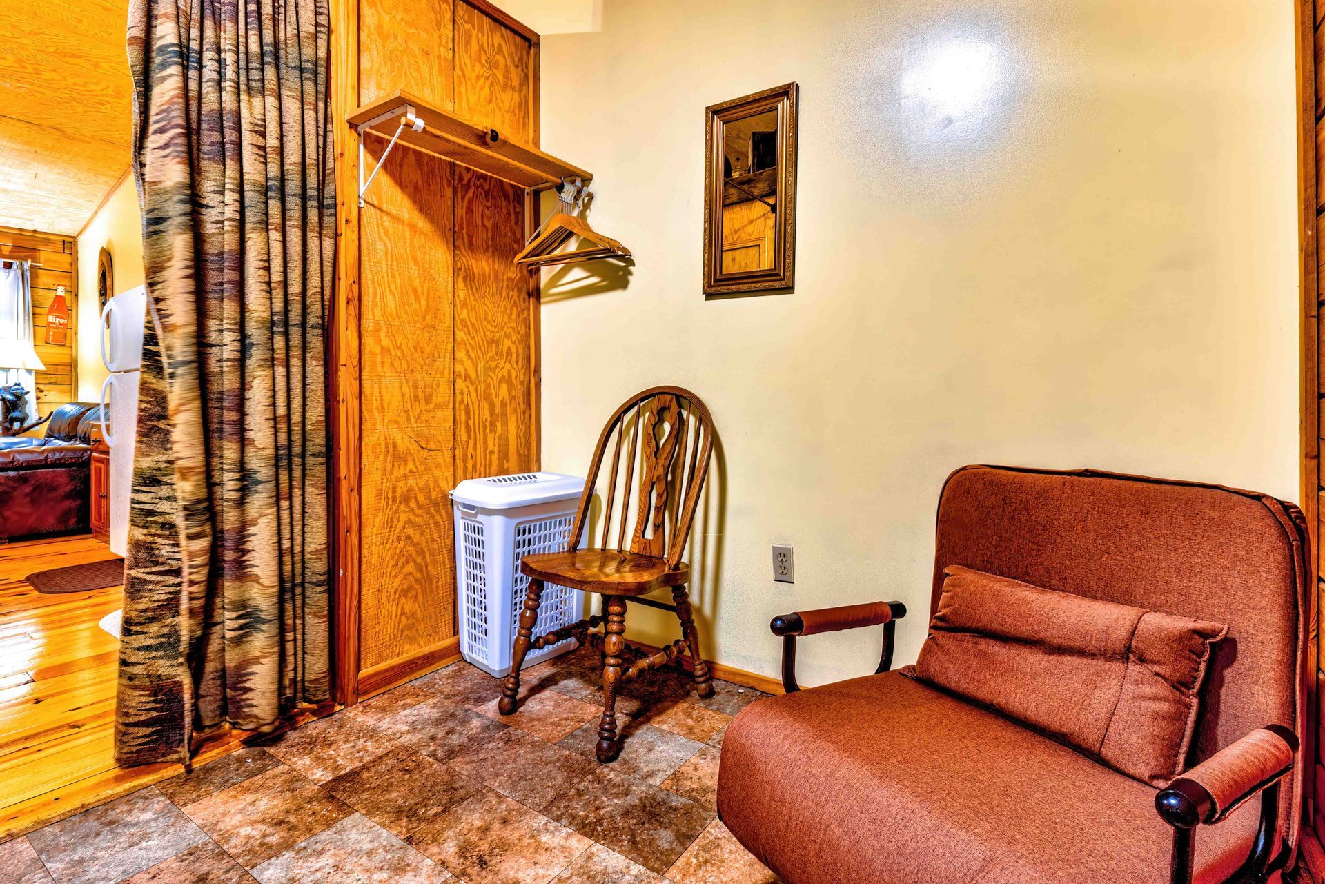 Small furnished room with a brown couch, wooden chair, coat rack, laundry basket, and patterned curtains.