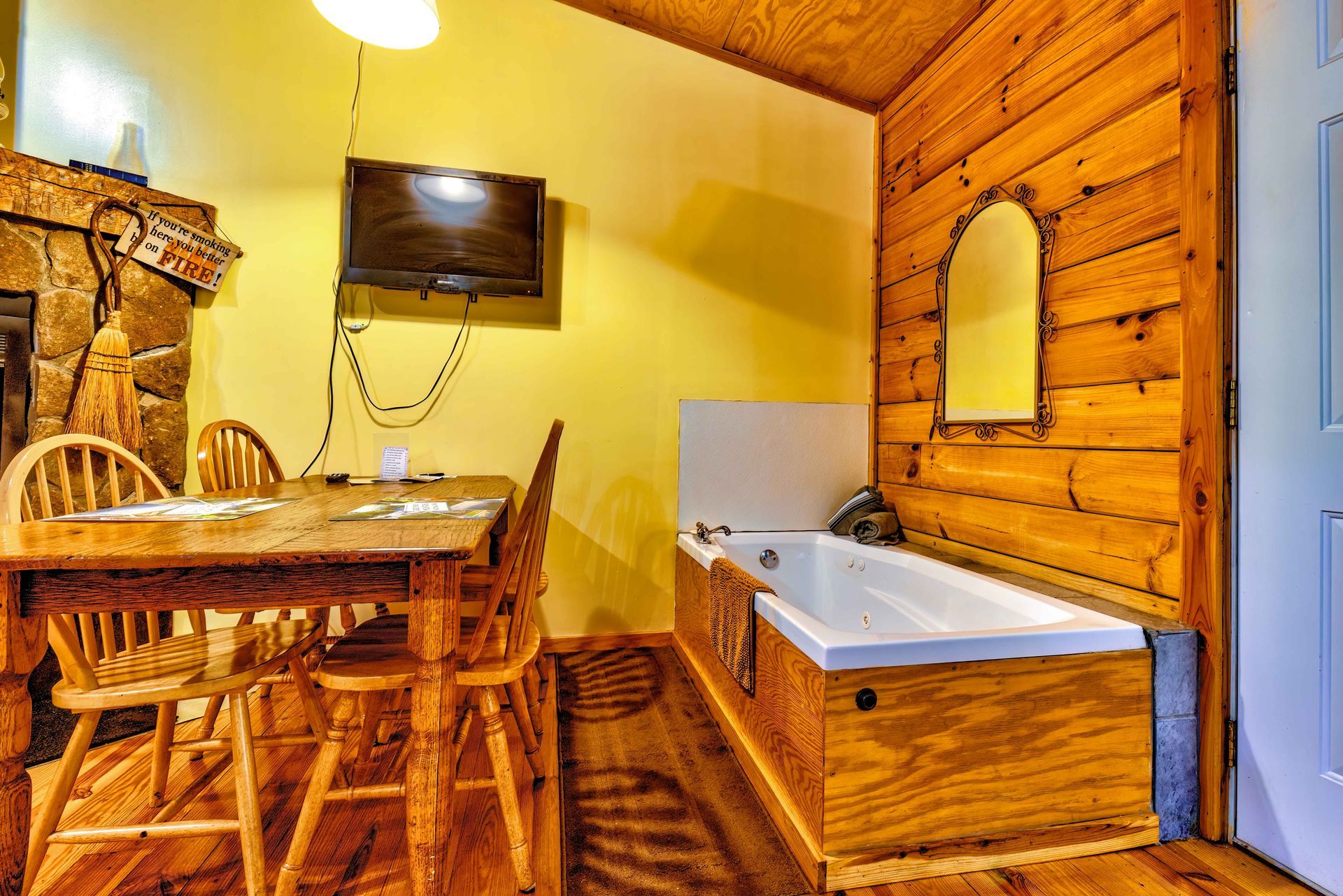 Rustic cabin kitchen with wooden dining table, chairs, stone fireplace, wall TV, and a bathtub along the wood-paneled wall