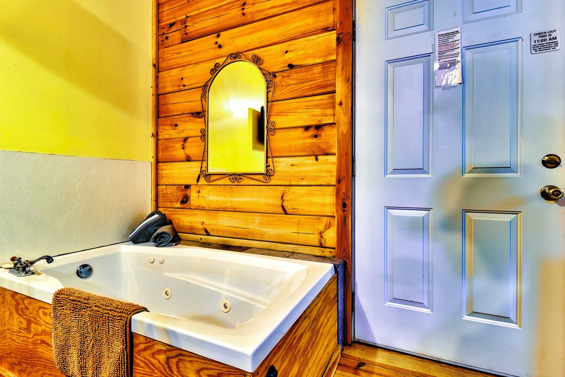 Bathroom with white soaking tub, wood-paneled wall, yellow accents, mirror, and closed white door.