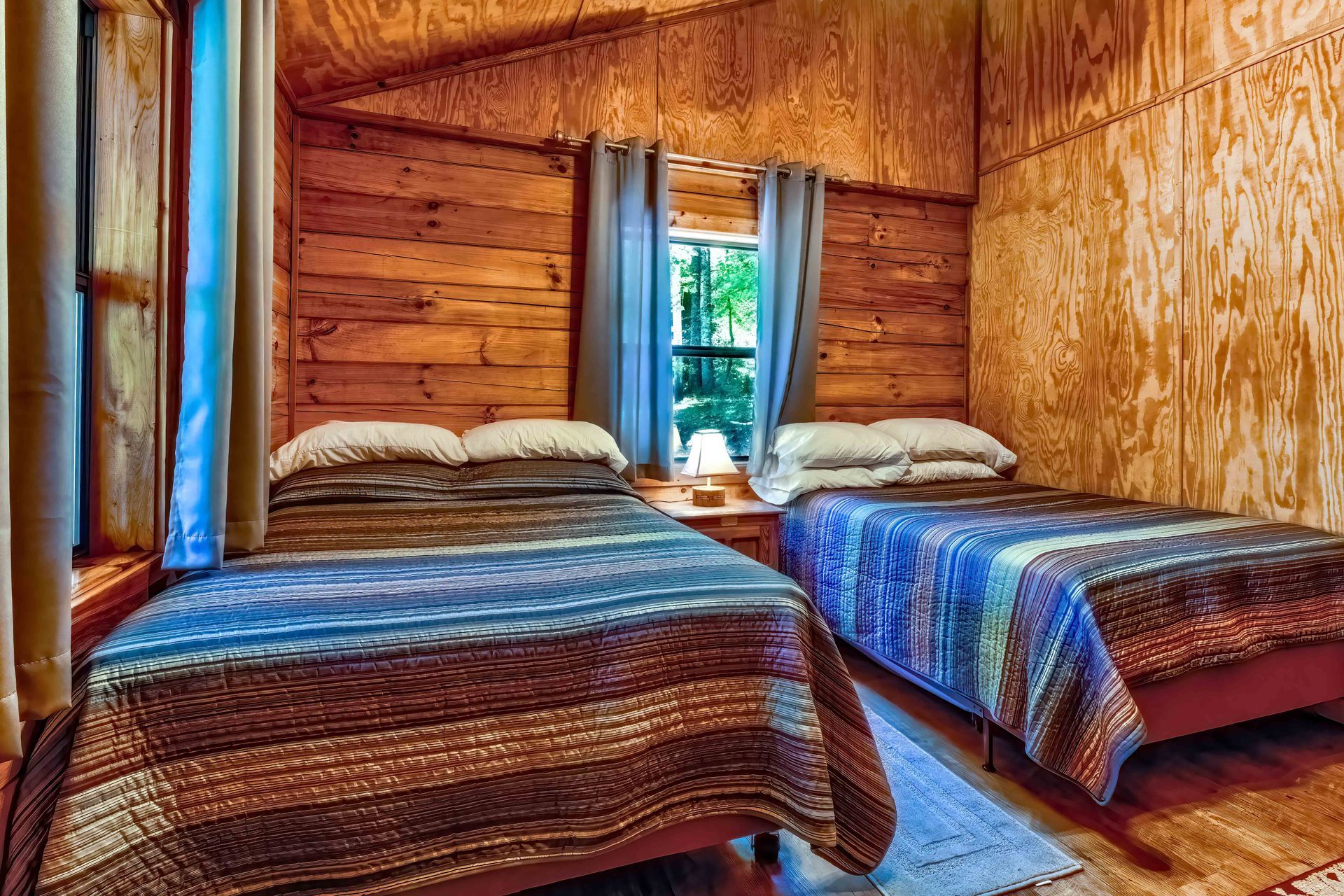 Two twin beds with patterned blankets in a rustic wood-paneled cabin room, sunlight through the window.