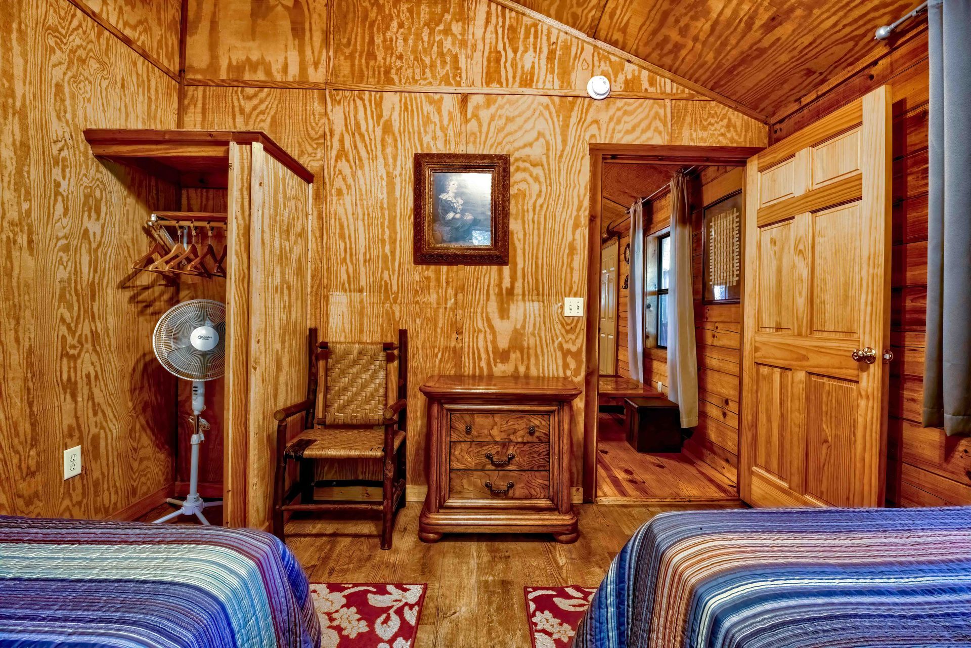Wood-paneled cabin room with twin beds, a chair, fan, and small table under a framed picture.