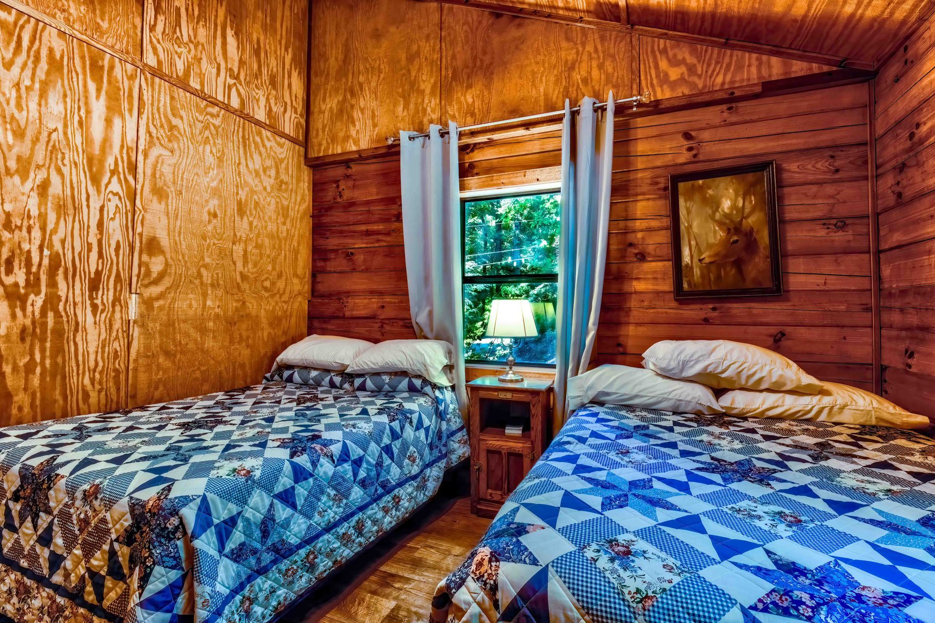 Cozy wooden cabin bedroom with two twin beds, blue quilts, and a window with curtains.
