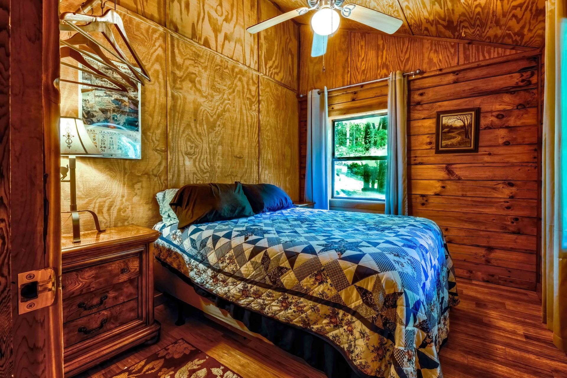 Cozy rustic bedroom with wood-paneled walls, blue quilted bed, window, and ceiling fan