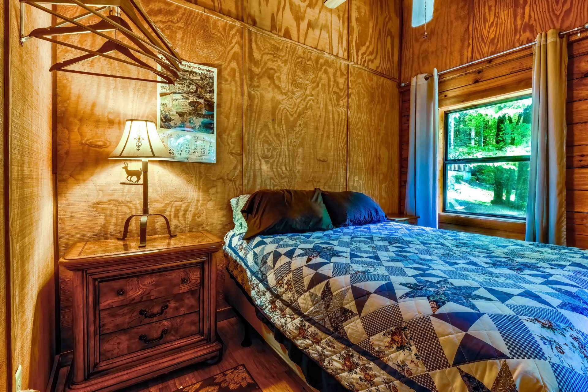 Cozy bedroom with quilted bed, wooden walls, nightstand lamp, and bright window view