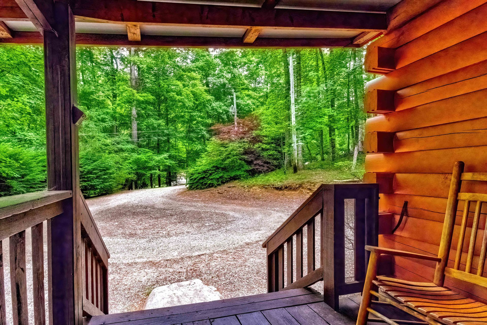 Covered cabin porch overlooking a gravel drive and lush green forest.