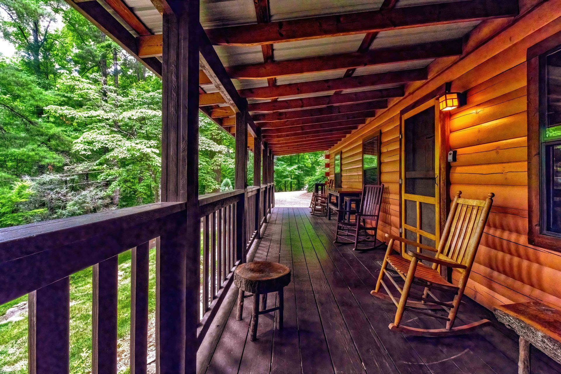 Covered log cabin porch with rocking chairs and stools overlooking a green forest