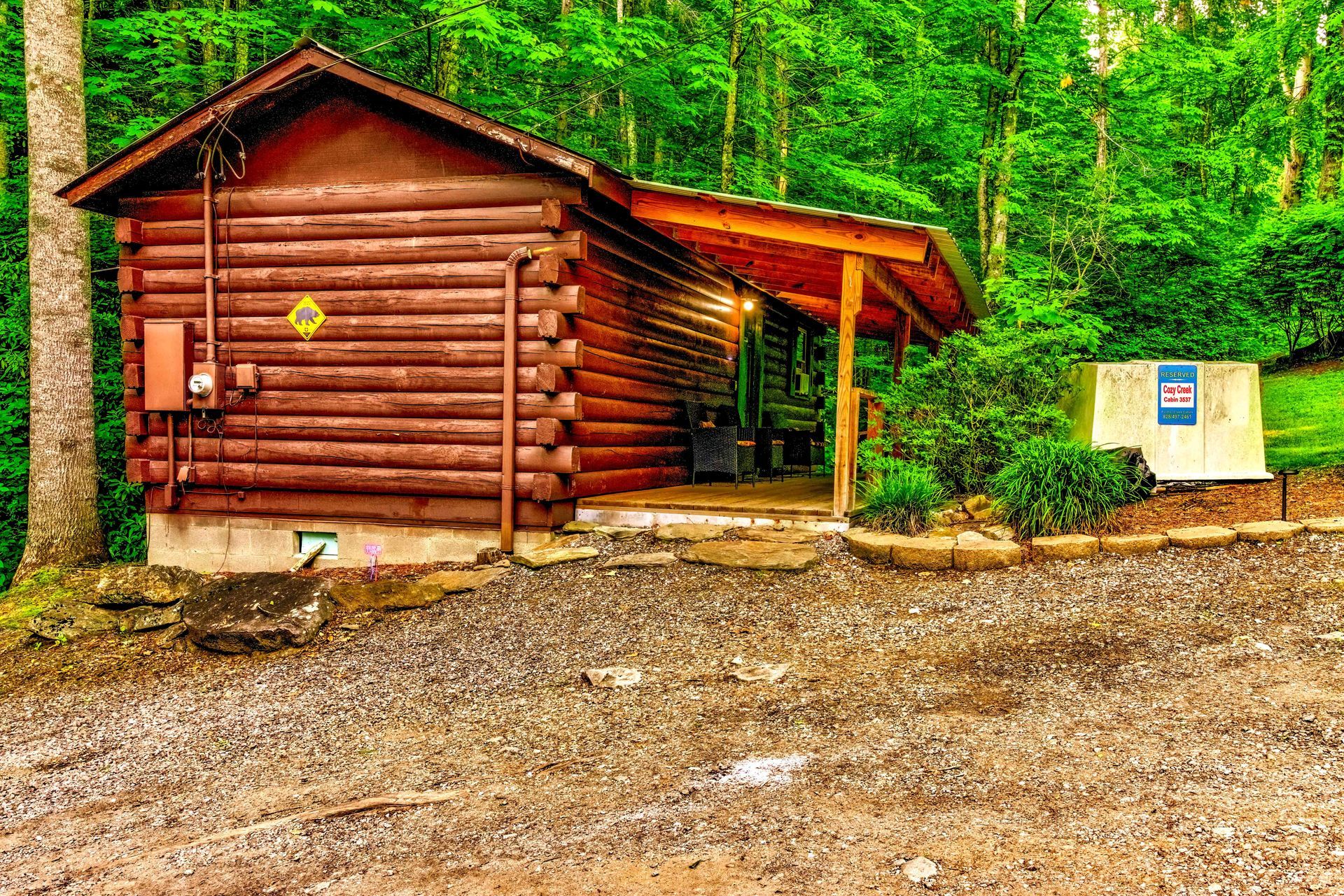 Rustic wooden cabin beside a gravel clearing, surrounded by lush green forest.