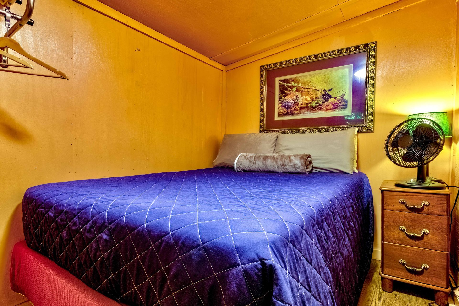 Cozy bedroom with a purple bedspread, yellow walls, framed artwork, and a small fan on a nightstand.