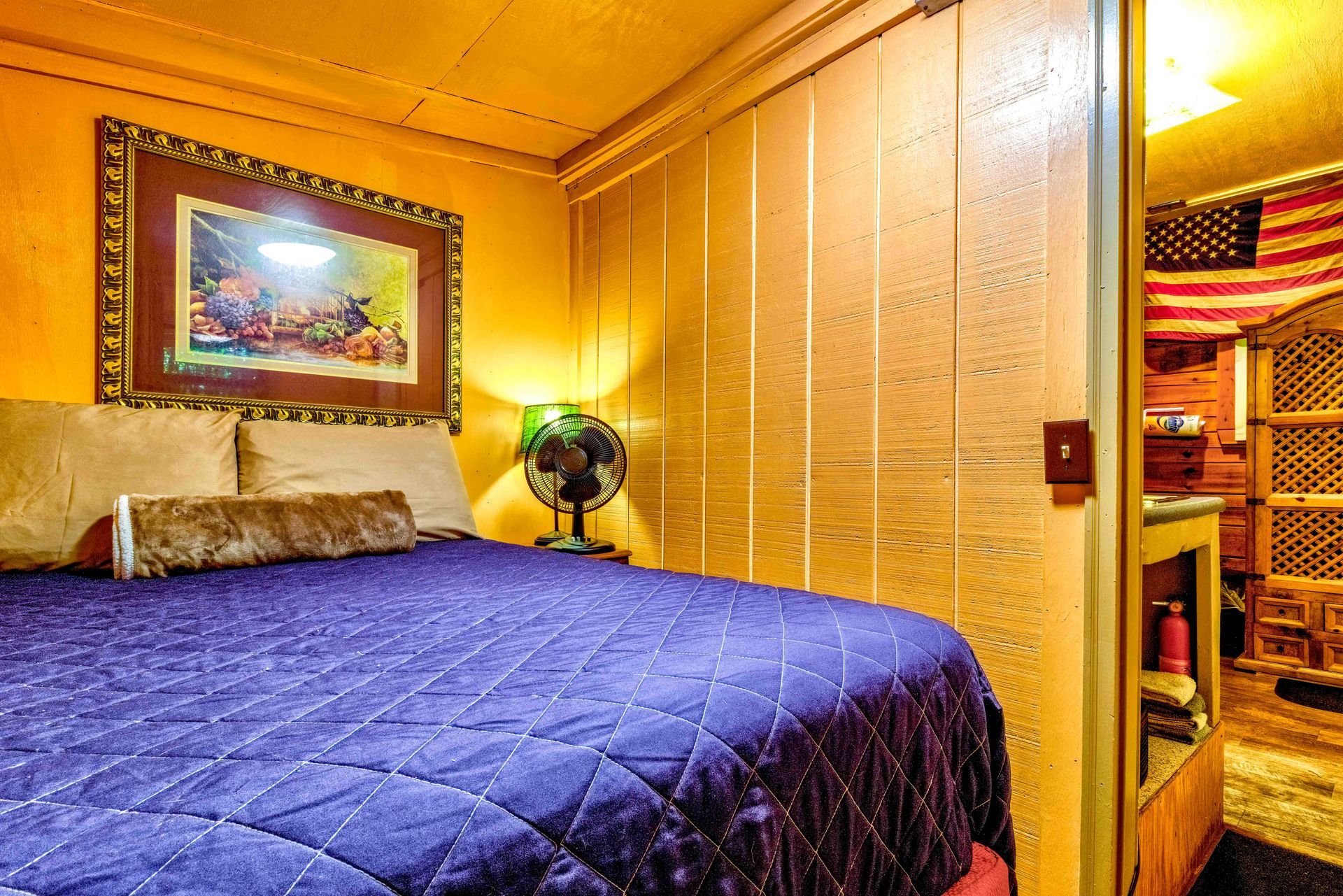 Small bedroom with a blue bedspread, framed wall art, fan, and American flag decor