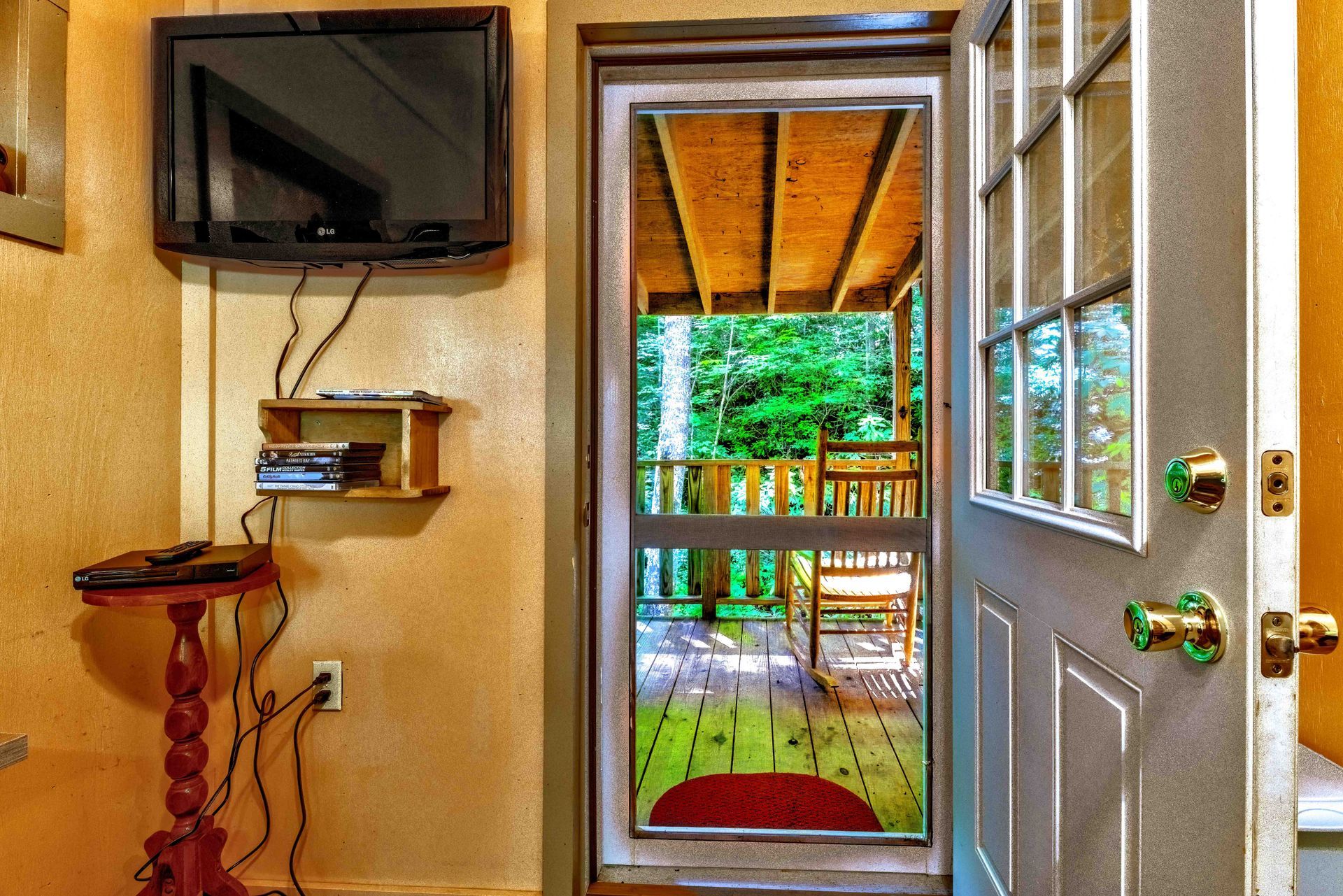 Small room with TV, wall shelf, and open door leading to a porch and green yard.