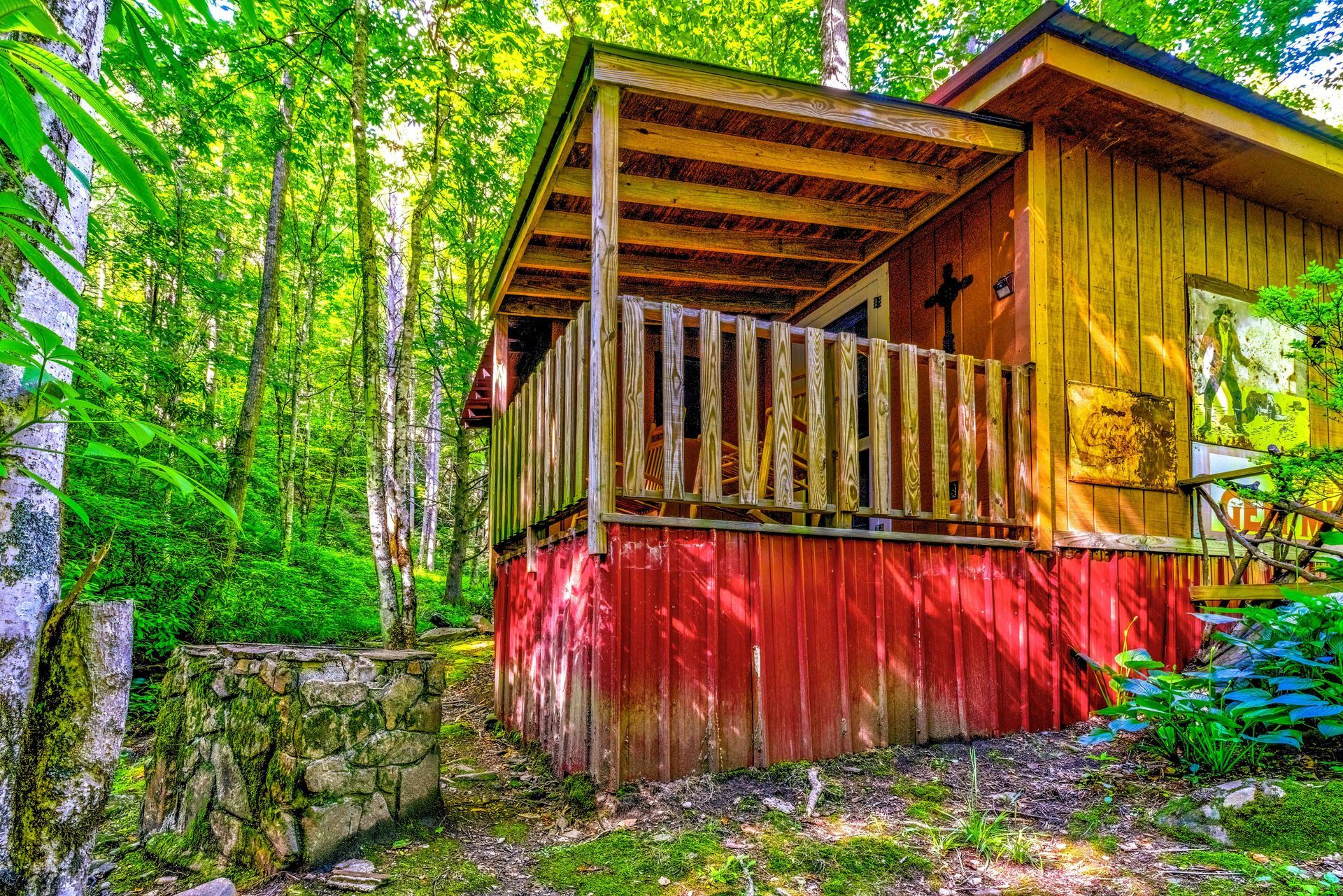 Rustic wooden cabin with red lower siding beside a stone wall in a lush green forest