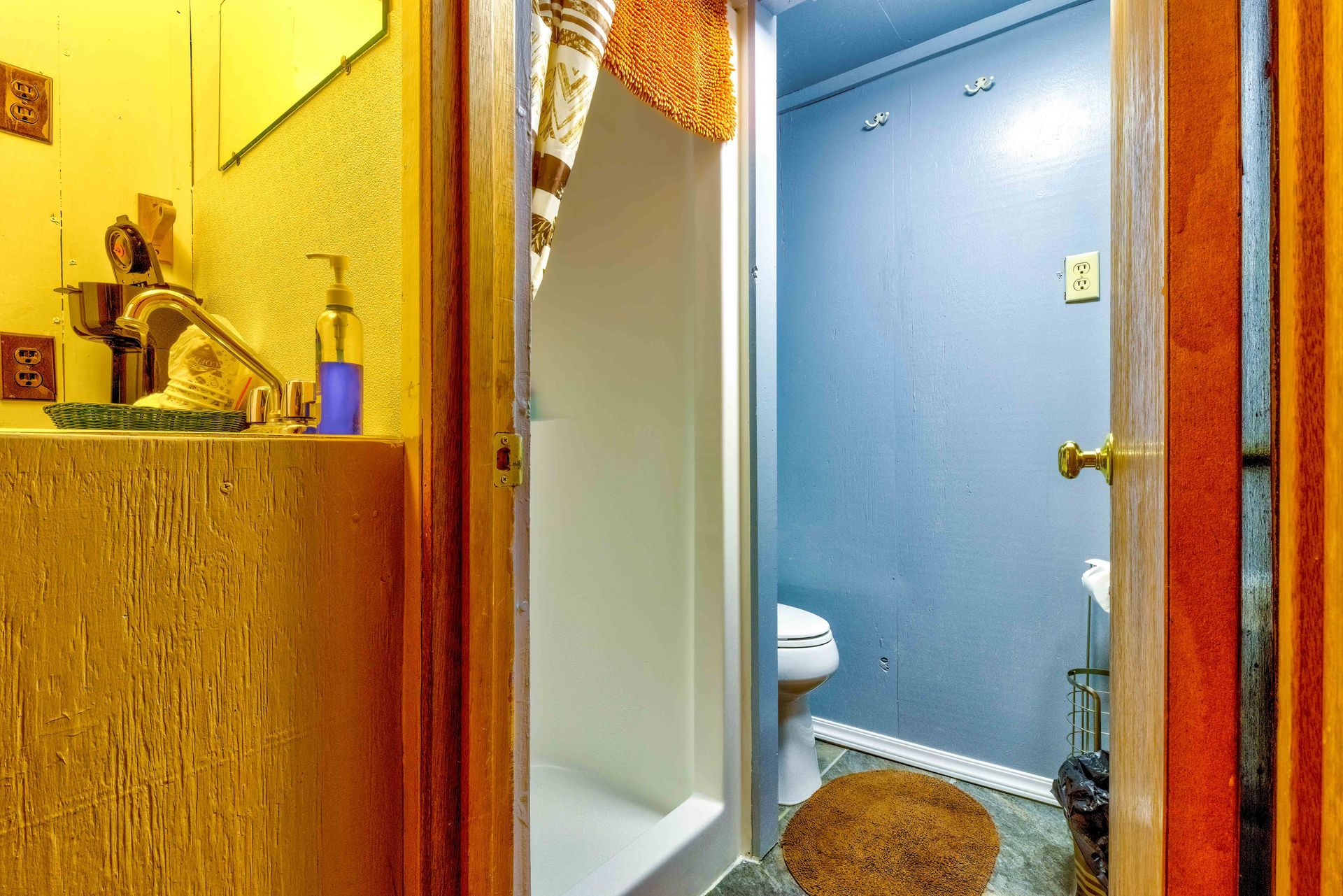 Open bathroom door showing a blue toilet room and a beige shower stall beside a sink.