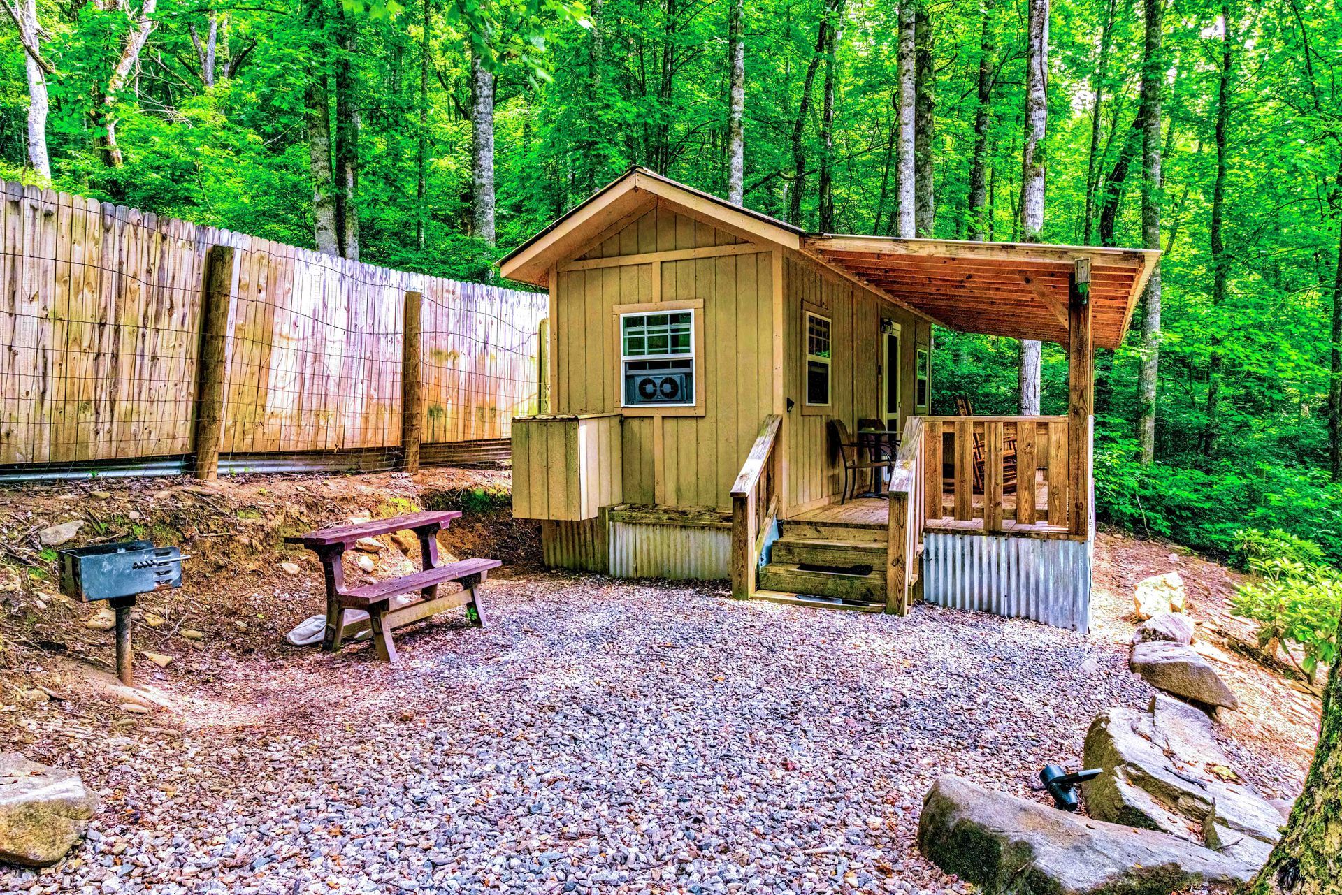 Small rustic cabin in a wooded area with a picnic table and gravel clearing