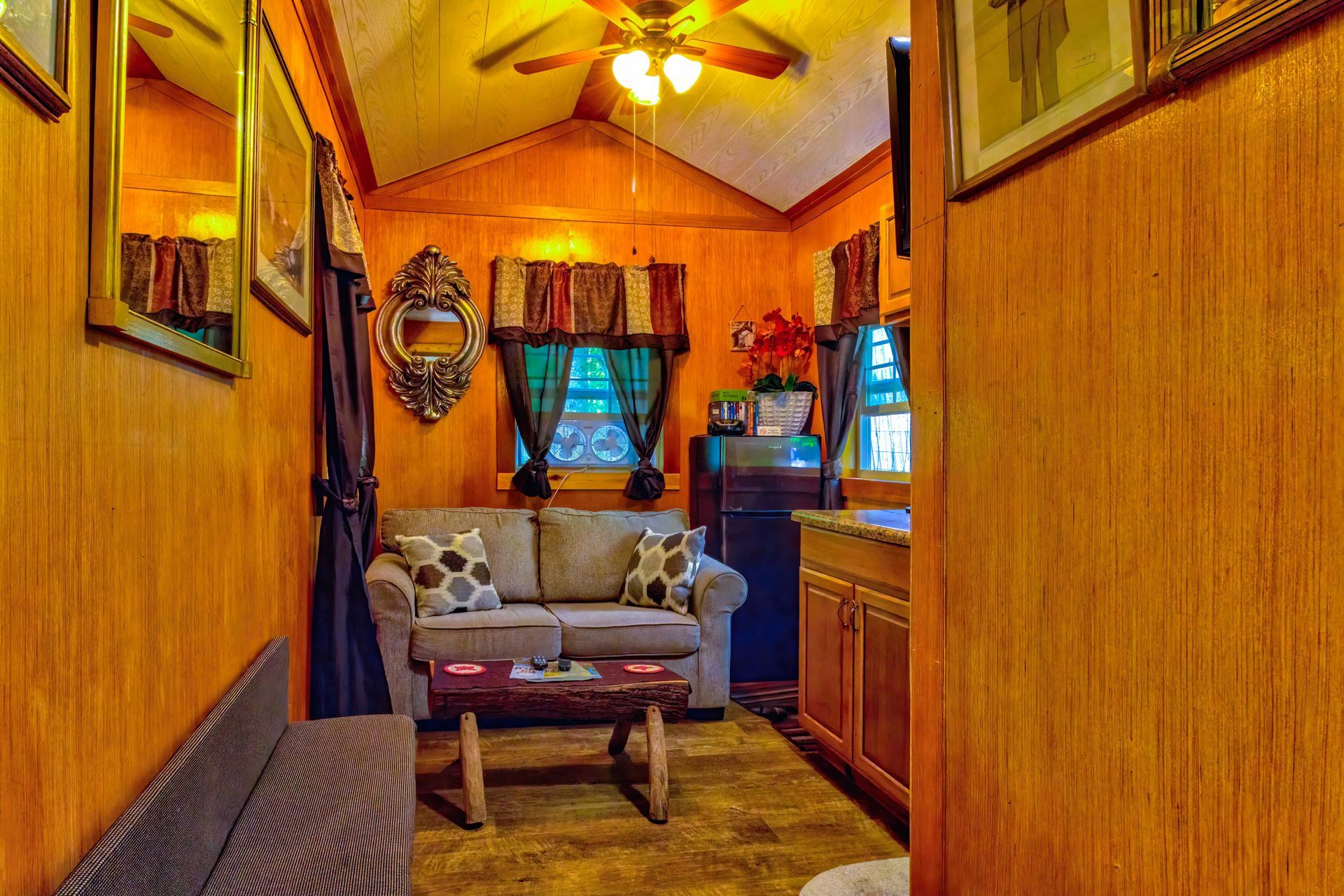Narrow wood-paneled room with a loveseat, ceiling fan, and a window with blue curtains.