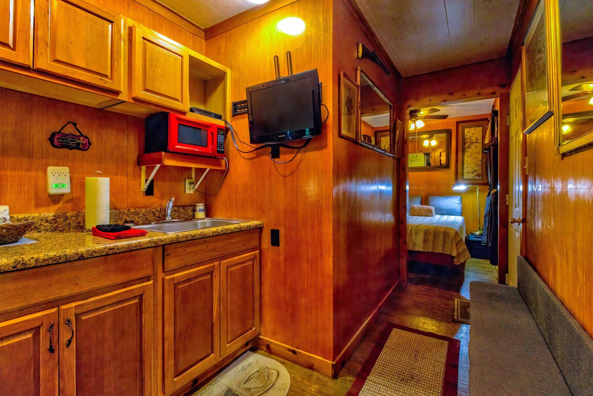 Wood-paneled RV kitchen and hallway with red cabinets, granite counter, and a small wall-mounted TV