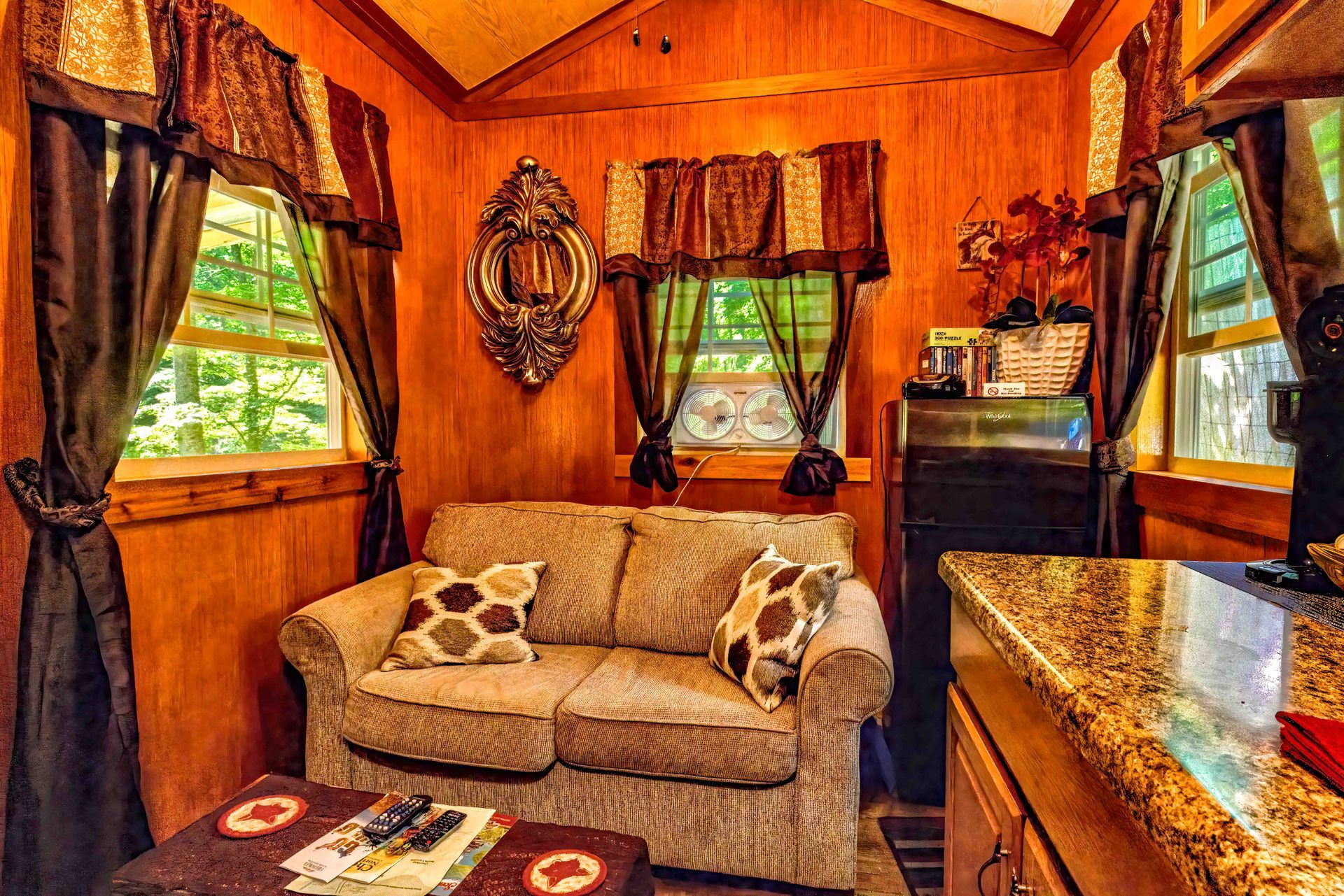 Warm wood-paneled cabin interior with a beige loveseat, patterned pillows, and windows with red curtains.
